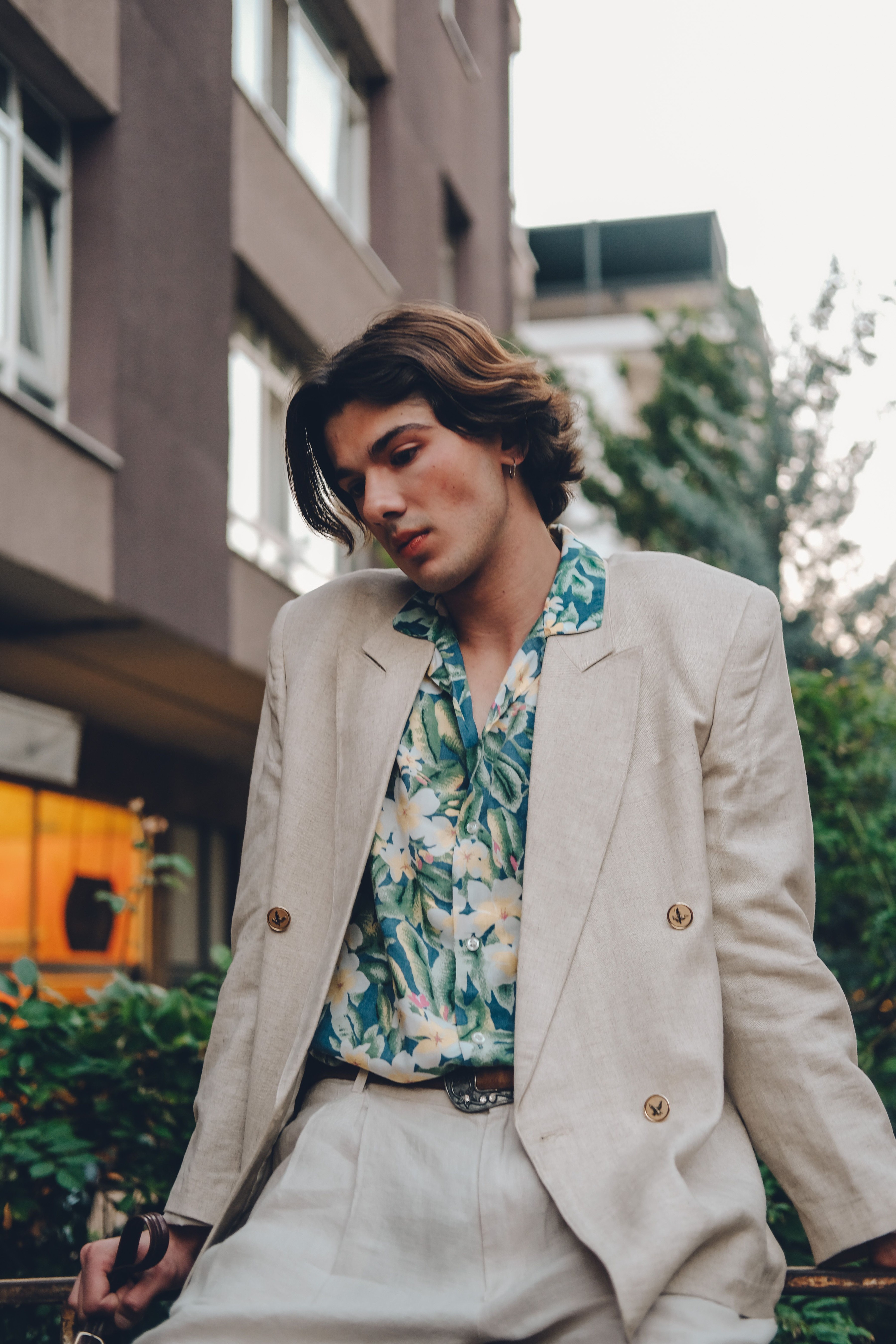 Homem elegante usando terno de linho bege com camisa floral, sentado ao ar livre em ambiente urbano