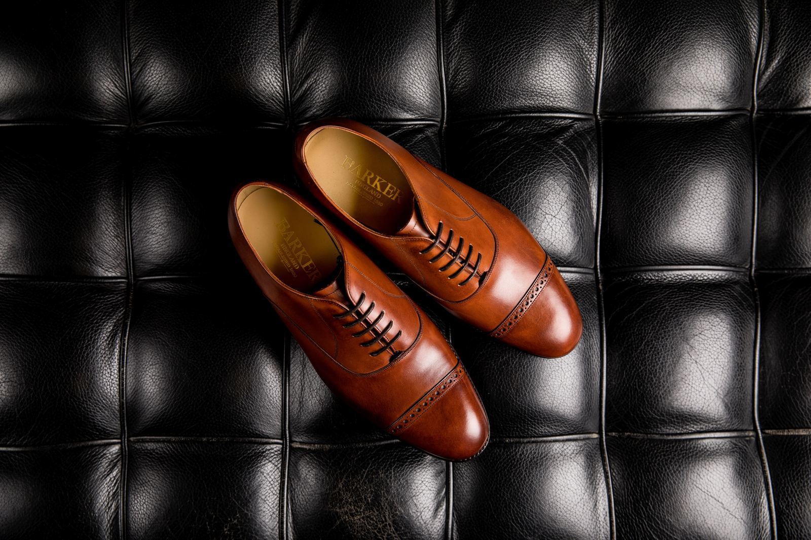 Par de sapatos Oxford de couro marrom sobre superfície escura, com destaque para os cadarços e o acabamento artesanal